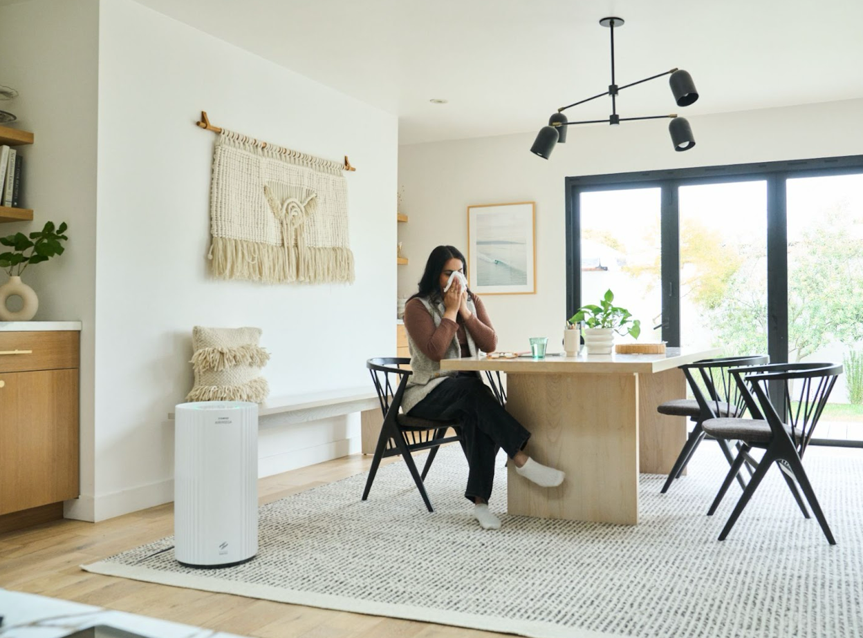 A person blowing their nose in their house next to an Airmega.